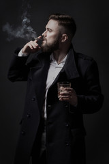 Attractive bearded man in a black coat smoking a cigar with pleasure. He holds a glass with some drink. He is stylish, strong and masculine, a cloud of cigar smoke rises above it.