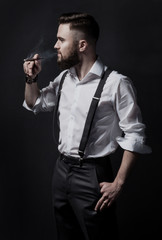 Young stylish bearded man in a white shirt and suspenders smoking a cigar in a dark room. He's a smart, educated, handsome and attractive.