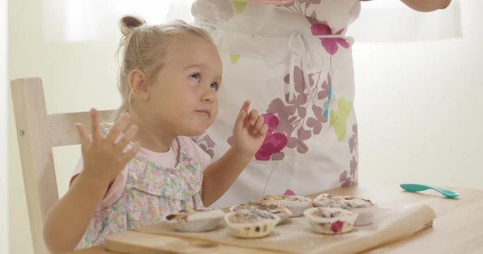 Adorable female toddler watching unidentifiable woman sift sugar as it falls on freshly baked muffins in wooden tray