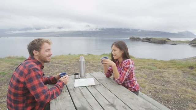 Couple camping sitting at table drinking coffee from thermos bottle flask by lake on Iceland. Campers woman and man relaxing taking break on road trip in beautiful Icelandic nature.