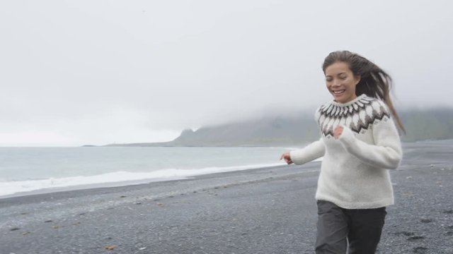 Happy woman having fun running on Iceland beach laughing joyful and playful wearing Icelandic sweater on black sand beach. Female model girl enjoying nature landscape at ocean sea. RED EPIC 90 FPS.