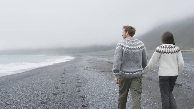 Couple walking on beach holding hands on Iceland. Romantic couple walking wearing Icelandic sweater on black sand beach from behind showing rear view of back. Tracking steadicam with RED EPIC 60 FPS.