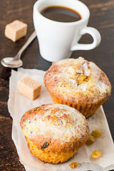 Dessert.    Cupcakes with raisins on parchment paper, a cup of coffee, sugar cubes and spoon on a brown wooden table.