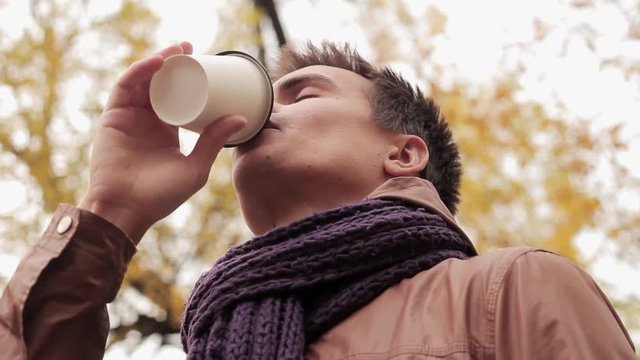 Young man in brown jacket and knitted scarf in autumn park drinks coffee out of paper cup close up