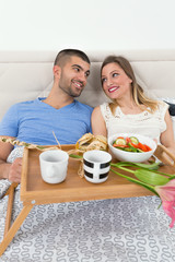 Obraz na płótnie Canvas Young couple is enjoying romantic meal in a hotel room. They are lying in bed and smiling to each other while holding a tray with a breakfast and bouquet of flowers.