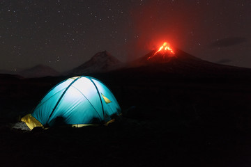Volcanic landscape of Kamchatka: night view of illuminated tourist tent on background eruption...