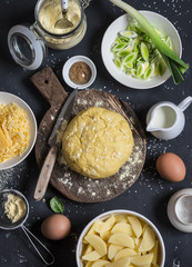 Baking ingredients - dough, potatoes, cheese, leek, cream. Cooking pie with leek, potato and cheese. On a dark background, top view