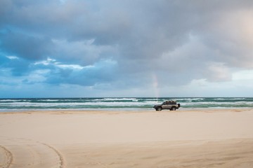 Fototapeta na wymiar Lonely car passes rainbow at sunset on Fraser Island coast