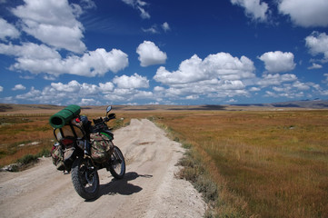 Enduro motorcycle traveler with suitcases standing on a dirt road vanishing into the skyline under a blue sky with white clouds, Plateau Ukok, Altai mountains, Siberia, Russia.