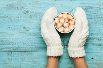 Wall murals Chocolate Woman hands in mittens hold cup of hot cocoa or chocolate with marshmallow on turquoise vintage table from above. Flat lay style.