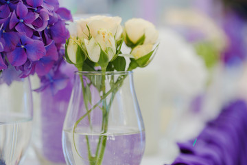 A vase with white rose buds stands behind a vase with violet lil