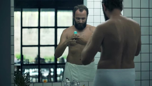 Young man in towel applying antiperspirant on his armpit in bathroom 
