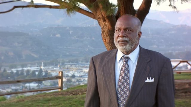 African American Older Mature Male Walks Forward Outside By A Tree And Smiles