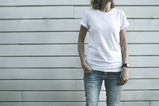 Front view. Girl in a white T-shirt and blue faded jeans standing with her hand in her pocket, on the other hand, the smartwatch. On background a white wall of wooden planks.Mock up.