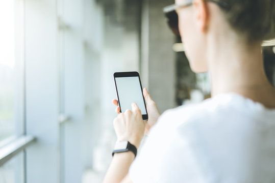 Mock up.Back view,girl standing in a room near a window and using digital gadget.Woman,dressed in white t-shirt,holding smartphone in her hand and pressing finger on blank screen.Space for text.