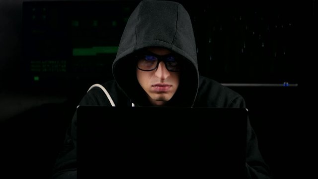 hacker with glasses and in a hooded working on a computer in a dark office room