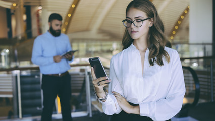 Beautiful young business woman standing in the lobby, holding a smartphone. In the background is a businessman who checks email on the tablet computer. Blurred background.