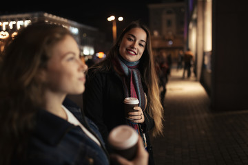 Two women friends walking in city street at night and drinking coffee