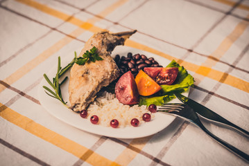 Gourmet roasted rabbit leg with rice and beans, white sauce. Meal is served on white simply plate and checked table cloth, with cutlery. Health and light meal good for diet.