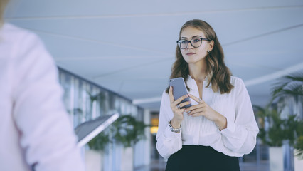 Beautiful young business woman with dark hair and glasses standing in the lobby and holding a smartphone . In the foreground is a business woman with a tablet computer. Blurred foreground .