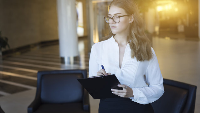 Business woman standing in a lobby and takes notes. Young girl in a white blouse standing in the lobby, and holding the clipboard. Blurred background. Solar effect.
