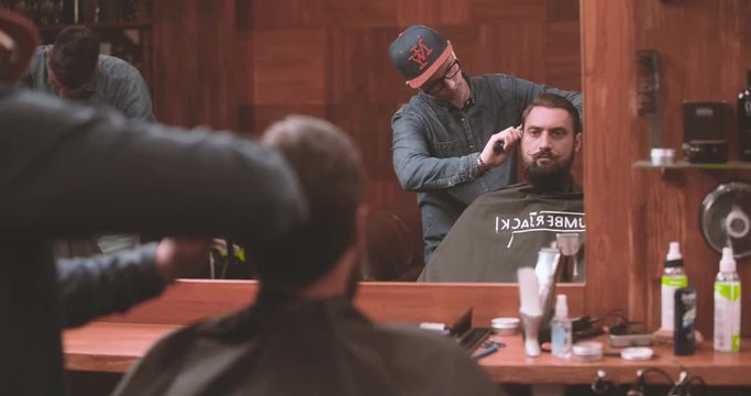 Hairdressing process at barbershop 4k video. Barber combing bearded lumberjack client head in salon mirror