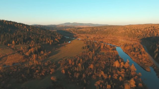 Shooting with quadrocopter. Aerial View of the mountain river. Country landscape at sunset. Smooth field, golden crown of the trees, a narrow river bed, rural road. The shadows of sunset.