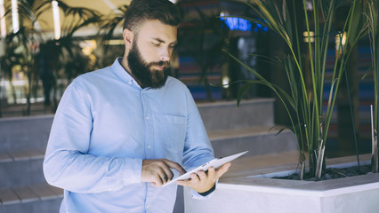 A bearded man with dark hair in a light blue shirt uses a tablet computer.Businessman checks mail on a digital tablet.Selective focus, film effect, blurred background.