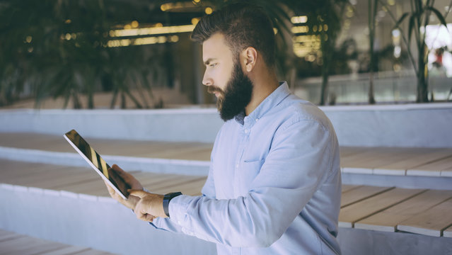 Hipster man sitting on bench and surf the Internet on tablet computer. Young bearded businessman in a blue shirt sitting and holding a digital tablet.