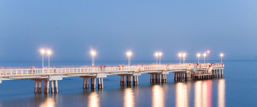Molo di Marina di Massa in Toscana durante il tramonto con luci che si riflettono sul mare calmo sotto al ponte d'estate.