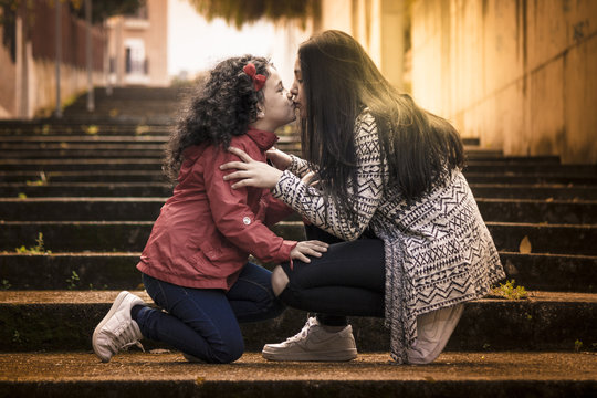 Ni&ntilde;a y adolescente, hermanas felices d&aacute;ndose un beso en las escaleras con el sol al fondo y a contraluz.
