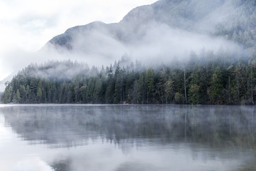 Buntzen Lake, Vancouver, British Columbia, Canada. Foggy lake in the morning. Evergreen forest reflection and mountain background.