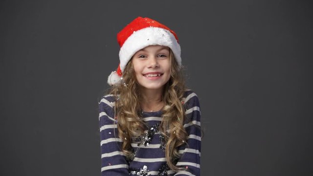 Cute little girl in a christmas cap throwing up confetti, gray background