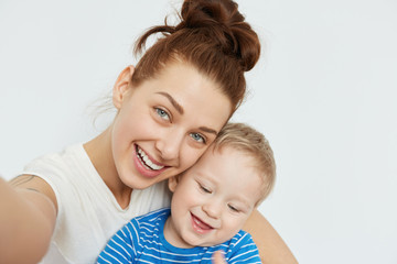 Positive family selfie with young mommy and toothless kid smiling together on white background. Playful state of mind and happy mood of attractive woman makes this shot fabulous, heartwarming.
