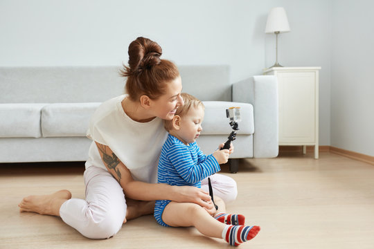 Sideways portrait of happy young Caucasian mother and son sitting at the floor at home, making selfie. Smiling woman in white clothes hugging her baby. Kid taking a picture with selfie stick.