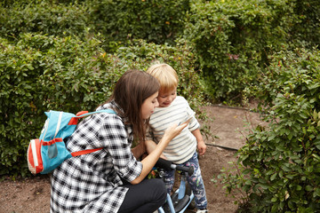 Beautiful shot of caring mother walking with her son outside. Young Caucasian woman showing photos to her baby on digital device. Pretty mom in checked shirt with childish rucksack sitting on hunkers.