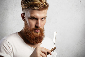 Headshot of attractive professional barber with thick red beard and mustache, holding razor. Young Caucasian hairdresser wearing white t-shirt standing in studio with cut-throat razor in his hands - obrazy, fototapety, plakaty