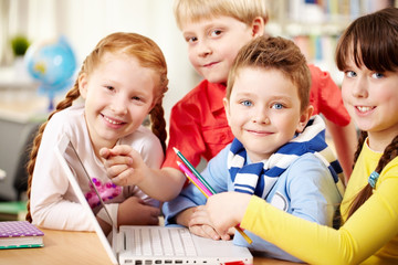 Obraz premium Four smiling schoolchildren sitting in classroom with a laptop, looking at camera and smiling