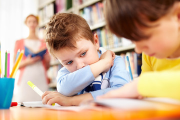 A little schoolboy sitting at table and looking at his working classmate