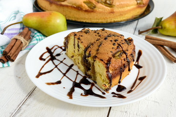 A piece of home baked biscuit flavored pear charlotte cake with cinnamon and chocolate syrup. On a white plate, on a light wooden background.
