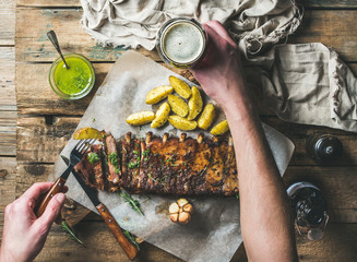 Man eating roasted pork ribs with garlic, rosemary and green herb sauce on rustic wooden table. Man' s hands holding fork with fried potato and glass of dark beer, top view