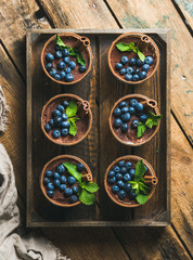 Homemade Tiramisu dessert in glasses with cinnamon sticks, mint leaves and fresh blueberries served in wooden tray over rustic wooden background, top view, vertical composition