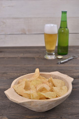Gold salted potato chips in a wooden bowl on a rustic brown table with a beer glass and a bottle in the background