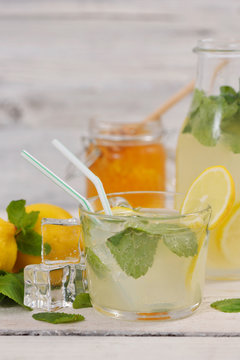 Glass of lemonade with lemon slice, mint leaves and ice cubes near lemons, lemonade bottle and honey jar on rustic white wood