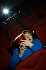 Naklejka premium Young man watching thriller at cinema hall and hiding behind popcorn bucket