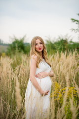 Fototapeta na wymiar Happy and yonge pregnant woman in a white dress with long blond hair in tall grass