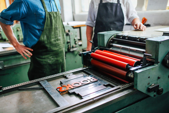 Senior craftsman/technician supervising young man on letterpress machine in book arts workshop, mid section