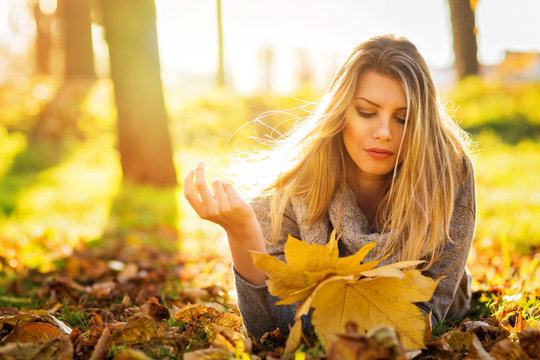 Beautiful young blonde woman relaxing in park in autumn holding leaves