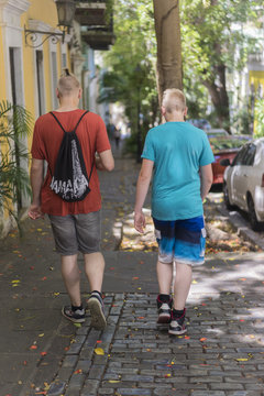 Two teenage brothers walking away from the camera along a cobbled sidewalk in a leafy urban suburb