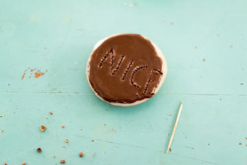 Chocolate cookie with the word Nice on it punched out with a wooden toothpick in the topping over a turquoise background with crumbs and copy space